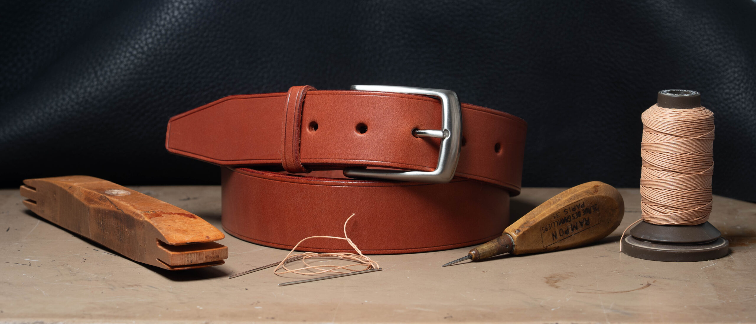 Ceinture en cuir cognac cousue main avec boucle laiton nickelé, entourée des outils de sellerie